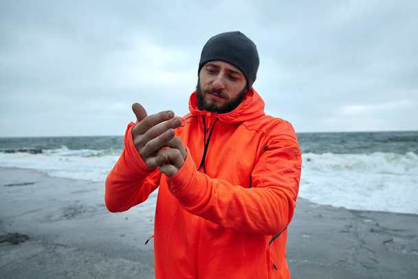 Image of attractive young athletic brunette male with beard standing over seaside on grey stormy weather and wearing warm sporty clothes, preparing for morning workout - Valokuva, kuva