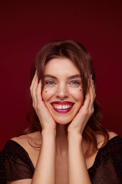 Charming blue-eyed young lady with brown wavy hair wearing silver stars on face and holding palms on her cheeks, smiling cheerfully to camera while posing against claret background - Foto, Imagem