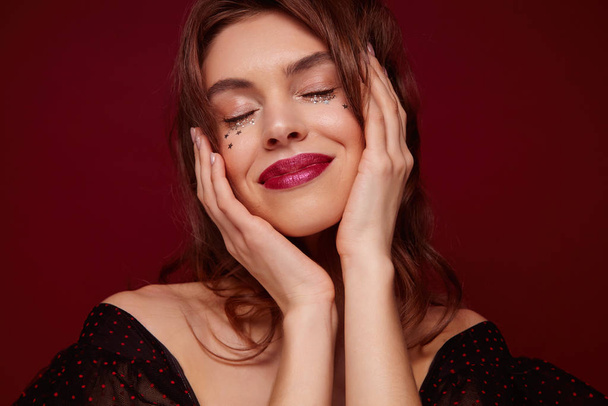 Enjoyable young brown haired pretty woman with red lips and little silver stars on her face smiling pleasantly with closed eyes, holding face with raised hands while posing against claret background - Fotó, kép