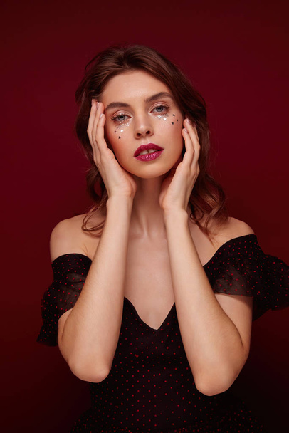 Studio photo of beautiful blue-eyed young brunette lady wearing festive makeup and elegant clothes over claret background, holding gently her face with raised hands and looking at camera - Photo, Image