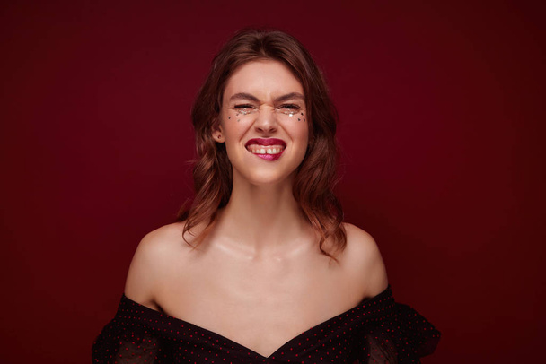 Portrait of young lovely positive female with brown hair frowning her face and biting underlip while looking at camera, dressed in elegant black top with red dots over claret background - Photo, Image