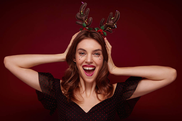 Joyful attractive young brunette woman holding hands on her head and looking happily to camera, preparing for christmas celebration and wearing holday hoop, isolated over claret background - Foto, Imagem