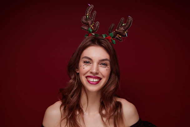 Close-up of cheerful young brown haired female with festive makeup wearing head horns while standing over claret background, smiling and expresses true positive emotions - Photo, Image