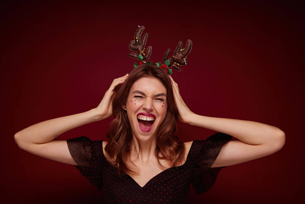 Overjoyed pretty young brunette woman with wavy hairstyle dressed in elegant clothes rejoicing while posing against claret background, wearing funny holiday hoop and laughing happily - Photo, Image