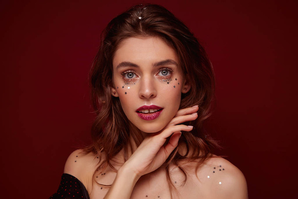 Studio photo of charming young brunette lady with burgundy lips and little silver stars on her face touching chin gently while looking at camera, isolated over claret background - Photo, Image