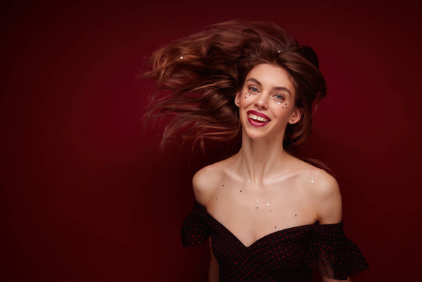 Portrait of pretty positive young long haired lady in black festive top with opened shoulders looking cheerfully at camera and waving her hair, isolated over claret background - Photo, Image