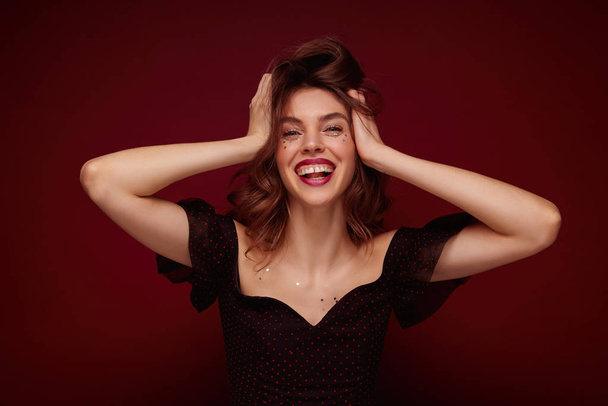 Indoor shot of young charming brunette lady in elegant black top with red dots clutching happily her head and laughing cheerfully while posing over claret background - Foto, afbeelding