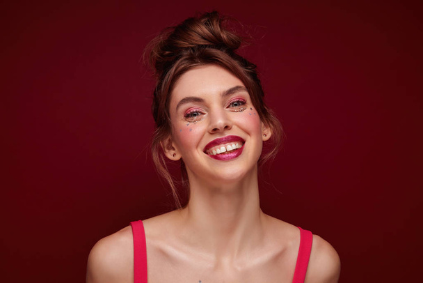 Porträt einer fröhlichen, schönen, brünetten jungen Frau mit Abendschminke, die ihr Haar im Dutt trägt, glücklich in die Kamera blickt und weit lächelt, isoliert vor weinrotem Hintergrund - Foto, Bild