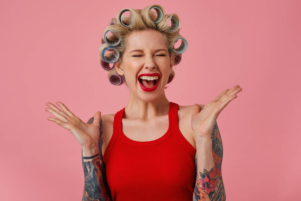 Overjoyed young blonde female with tattooes screaming happily with closed eyes and raising emotionally hands, having curlers on her head while posing over pink background - Fotó, kép