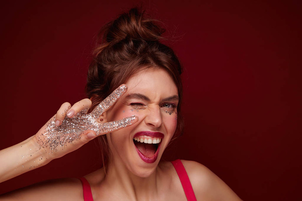 Joyful pretty young brunette female with claret lips wearing her hair in knot and raising victory sign with sparkles on fingers to her face, winking cheerfully at camera with wide mouth opened - Photo, Image