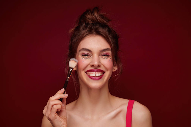 Happy young brown haired lovely woman with festive makeup wearing casual hairstyle while standing over burgundy background, meeting with friends in evening and being in high spirit - Foto, Imagem
