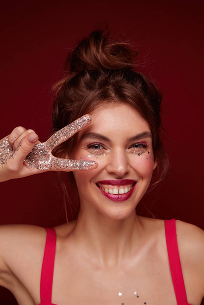 Cheerful young attractive brown haired lady going out for party and making festive makeup, smiling widely at camera and keeping raised fingers apart near her face, isolated over burgundy background - Photo, Image