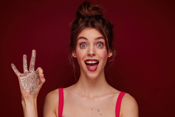 Open-eyed young pretty brunette female with bun hairstyle and festive makeup looking excitedly at camera and raising ok gesture with sparkles on her hand, isolated over burgundy background - Zdjęcie, obraz