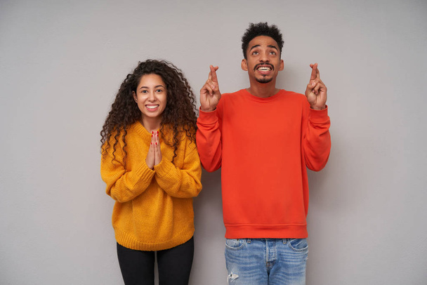 Studio shot of hoping attractive young dark skinned curly pair wearing woolen colourful sweaters and jeans while standing over grey background with raised hands and smiling widely - Valokuva, kuva