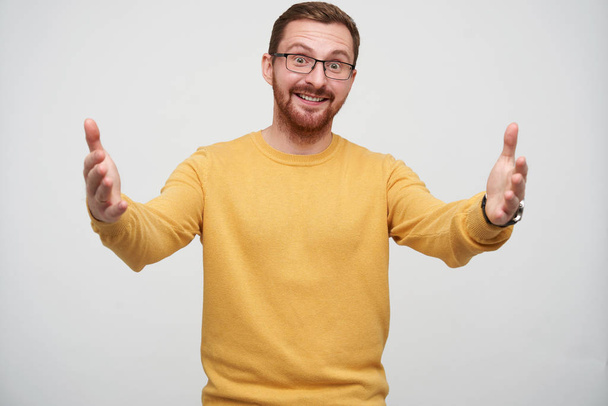 Cheerful young lovely brunette man in glasses with beard posing over white background with wide opened arms, looking positively at camera and smiling happily - Valokuva, kuva