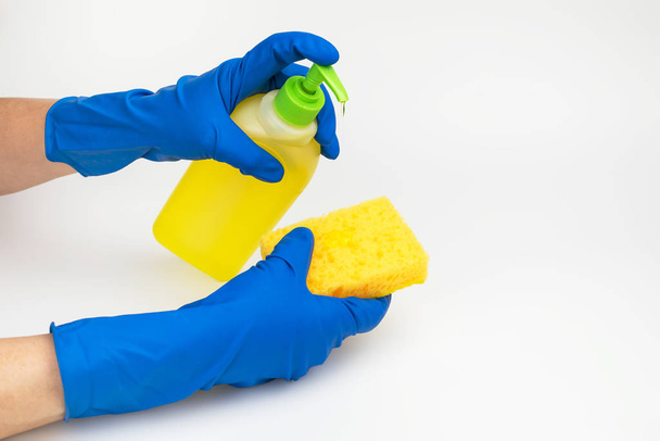 Handen in blauwe handschoenen giet wasmiddel uit de fles op de gele spons op een witte achtergrond - Foto, afbeelding