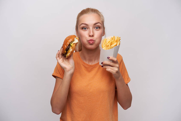 Portrait of charming pretty young blonde woman holding junk food and looking joyfully to camera, puffing out cheeks and being excited about tasty dinner, isolated over white background - Valokuva, kuva