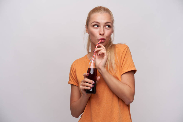 Pleasant looking young positive long haired blonde woman with casual hairstyle drinking soda with straw from glass bottle, dressed in orange t-shirt while standing over white background - Photo, image