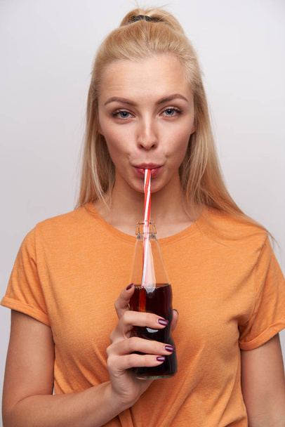 Piękna młoda blondynka z przypadkową fryzurą trzymająca butelkę z sodą i pijąca ją słomką, patrząca w kamerę ze spokojną twarzą, nosząca pomarańczowy t-shirt na białym tle - Zdjęcie, obraz