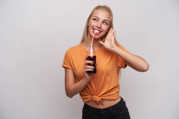 Indoor shot of joyful beautiful young long haired blonde female drinking soda and looking happily aside with wide smile, being in nice mood while posing against white background - Foto, immagini