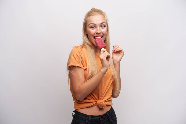 Indoor photo of happy lovely young long haired blonde woman with casual hairstyle looking cheerfully at camera while enjoying berry ice-cream in her hand, isolated over white background - Фото, изображение