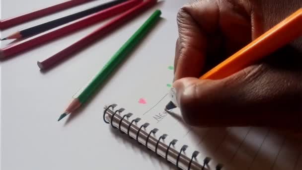 アフリカの手は、鋭いペンビデオでノートパッドに新年の決意の言葉を書いています。背景に色鉛筆がある. - 映像、動画