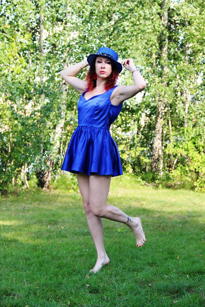 Women with red curly hair in a classic blue short dress and blue hat, jumping on one leg on the green lawn - 写真・画像