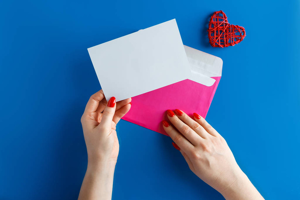 Roze envelop met een blanco kaart in handen op een blauwe achtergrond. Envelop met een schone wenskaart en een hart op een blauwe achtergrond. Concept voor ontwerp op Valentijnsdag. - Foto, afbeelding