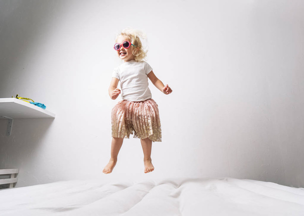 A little girl in pink sunglasses and a pink skirt jumps on a white mattress. - Фото, изображение
