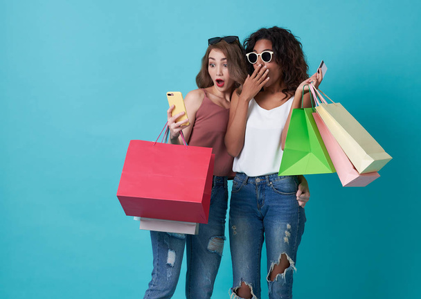 Portrait of two excited young woman hand holding mobile phone and shopping bag isolated over blue background. - Zdjęcie, obraz