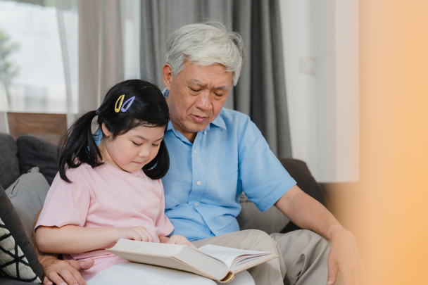 Aasialainen isoisä rentoutua kotona. Vanhempi kiinalainen, isoisä onnellinen rentoutua nuori tyttärentytär tyttö nauttia lukea kirjoja ja tehdä kotitehtäviä yhdessä olohuoneessa käsite. - Valokuva, kuva