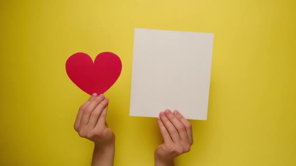 Vrouwelijke handen houden rood papieren hart en wit blanco door een gele papieren muur. Met plaats voor tekst. Valentijnsdag, Internationale Vrouwendag en verjaardagsconcept. - Video
