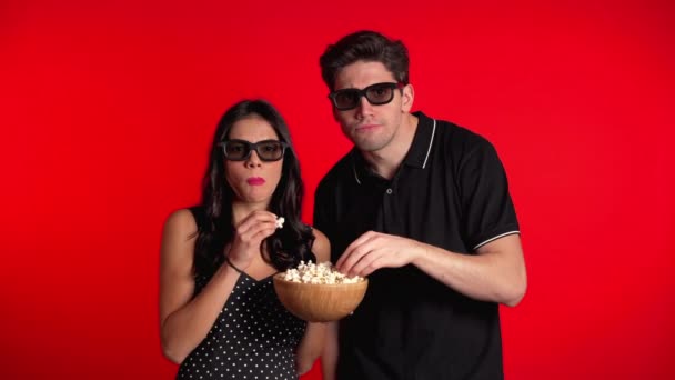 Young couple in cinema in 3d glasses watching fascinating horror movie and eating popcorn on red studio background. - Materiaali, video