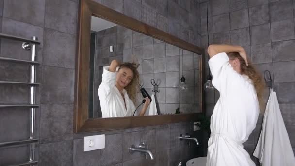 Vonzó szőke nő fehér fürdőköpenyben hosszú göndör hajszárító haj előtt tükör fürdőszobában otthon - Felvétel, videó