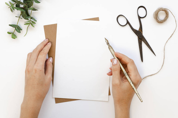 handgemachter Bohemien-Arbeitsplatz. leere Papierkarte und weibliche Hände halten Tintenstift bereit, um Schriftzug Grußtext zu schreiben. Eukalyptus, Schere und Bindfaden auf einem weißen Tisch - Foto, Bild