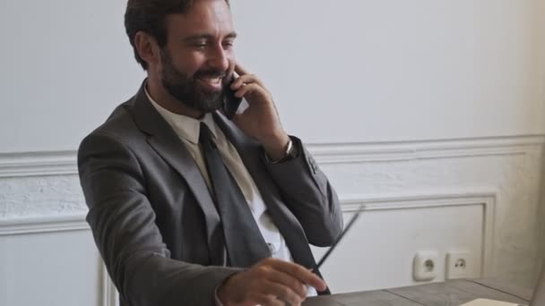 Cheerful handsome bearded businessman talking by smartphone while sitting at office - Materiał filmowy, wideo