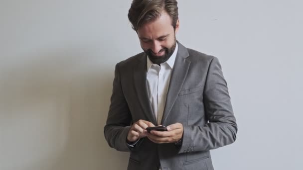 Happy handsome bearded businessman using smartphone while standing near the wall at office - Кадры, видео