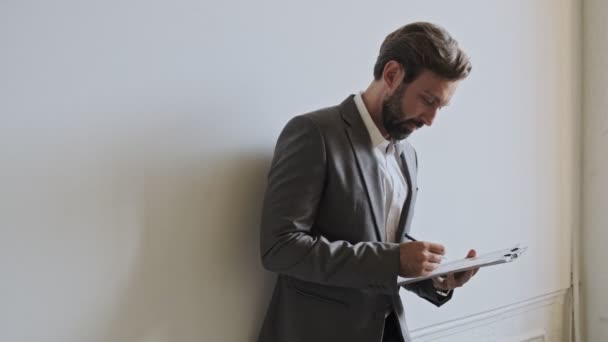 Concentrated handsome bearded businessman holding clipboard and checking something while standing near the wall at office - Filmagem, Vídeo