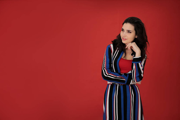 portrait of a beautiful girl with dark hair in a striped dress on a red background in a good positive mood, a young woman free space for notes - Photo, Image