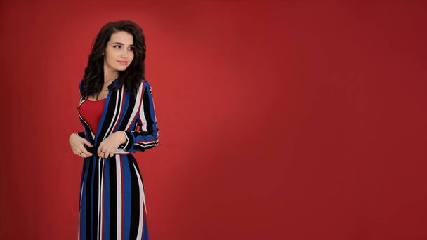 portrait of a beautiful girl with dark hair in a striped dress on a red background in a good positive mood, a young woman free space for notes - 写真・画像