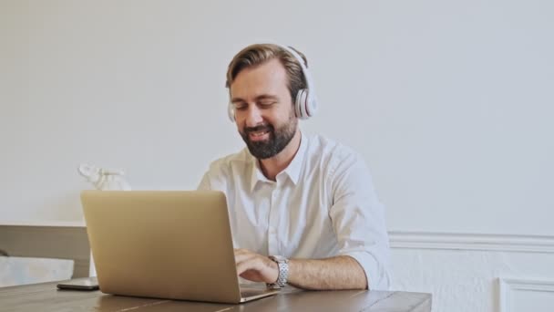 Smiling handsome bearded businessman listening music and using laptop computer while sitting by the table at office - Кадры, видео
