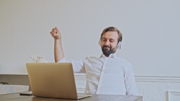 Cheerful handsome bearded businessman in headphones having fun and using laptop computer while sitting by the table at office - Video, Çekim