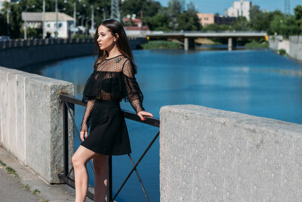 beautiful brunette stands on the bridge, near the river, in the city. a girl in a black dress puts a face to the sun and the wind that blows on her - Φωτογραφία, εικόνα