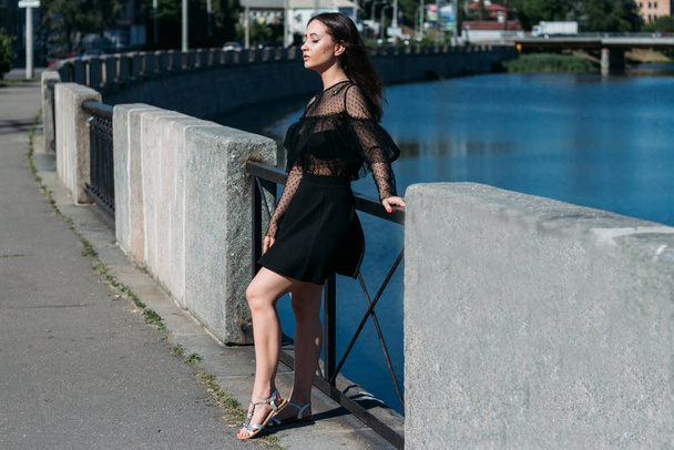 beautiful brunette stands on the bridge, near the river, in the city. a girl in a black dress puts a face to the sun and the wind that blows on her - Zdjęcie, obraz