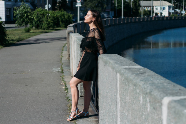 beautiful brunette stands on the bridge, near the river, in the city. a girl in a black dress puts a face to the sun and the wind that blows on her - Photo, image