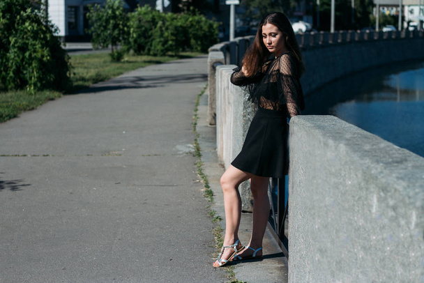 beautiful brunette stands on the bridge, near the river, in the city. a girl in a black dress puts a face to the sun and the wind that blows on her - Fotografie, Obrázek