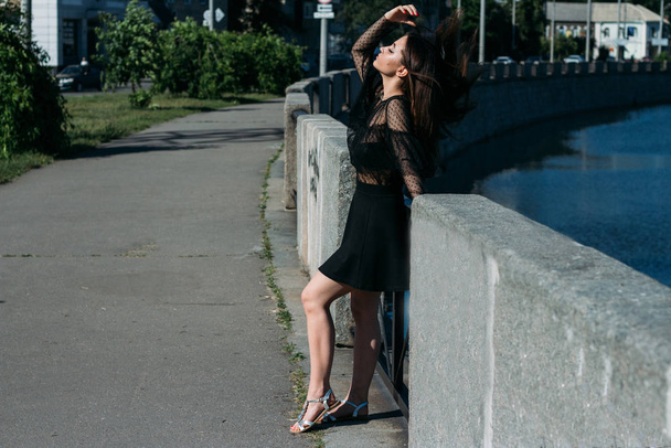 beautiful brunette stands on the bridge, near the river, in the city. a girl in a black dress puts a face to the sun and the wind that blows on her - Fotografie, Obrázek