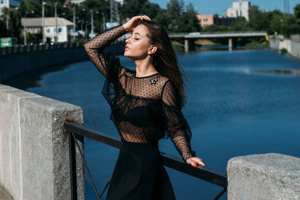 όμορφη μελαχρινή στέκεται στη γέφυρα, κοντά στο ποτάμι, στην πόλη. Ένα κορίτσι με μαύρο φόρεμα βάζει ένα πρόσωπο στον ήλιο και τον άνεμο που φυσάει πάνω της - Φωτογραφία, εικόνα