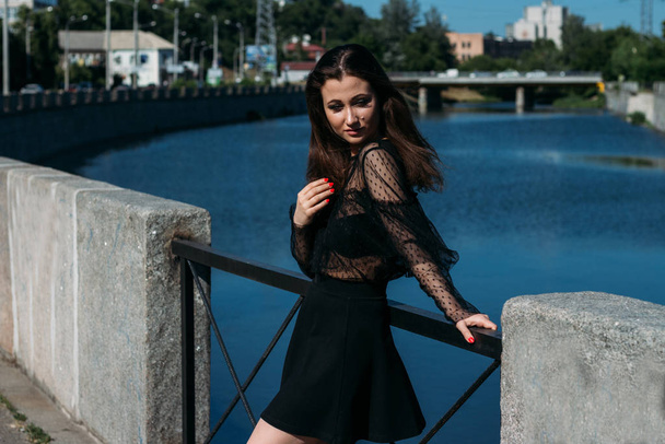beautiful brunette stands on the bridge, near the river, in the city. a girl in a black dress puts a face to the sun and the wind that blows on her - Fotoğraf, Görsel