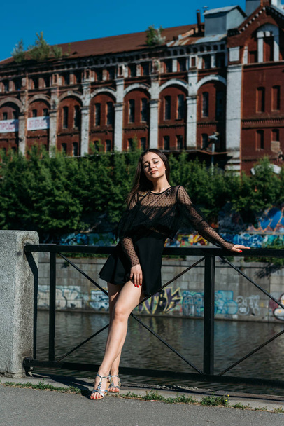 beautiful brunette stands on the bridge, near the river, in the city. a girl in a black dress puts a face to the sun and the wind that blows on her - 写真・画像